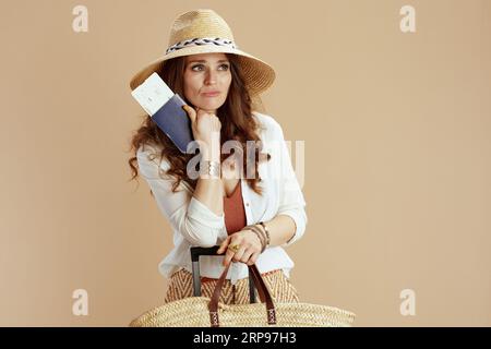 Vacanza al mare. donna triste e alla moda con blusa bianca e pantaloncini corti su sfondo beige con sacchetto di paglia, biglietto aereo, passaporto, trolley e cappello di paglia. Foto Stock