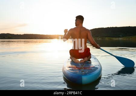 Uomo che pratica il paddle board su SUP board nel fiume al tramonto, vista posteriore Foto Stock