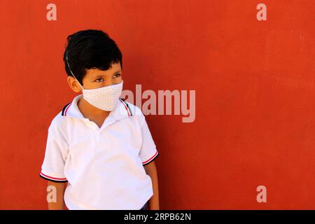 Felice e penoso Latino 8 anni ragazzo che indossa camicia bianca uniforme scolastica e maschera viso per la protezione nella pandemia di Covid-19 al ritorno a scuola Foto Stock