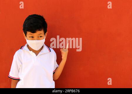 Felice e penoso Latino 8 anni ragazzo che indossa camicia bianca uniforme scolastica e maschera viso per la protezione nella pandemia di Covid-19 al ritorno a scuola Foto Stock