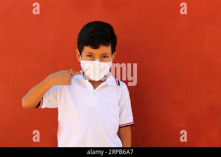 Felice e penoso Latino 8 anni ragazzo che indossa camicia bianca uniforme scolastica e maschera viso per la protezione nella pandemia di Covid-19 al ritorno a scuola Foto Stock