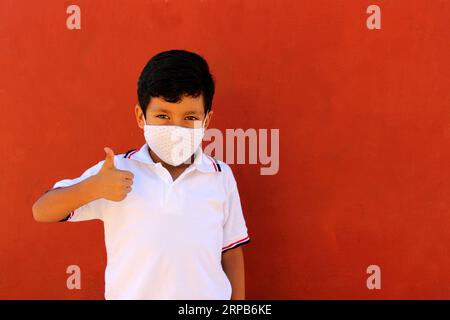 Felice e penoso Latino 8 anni ragazzo che indossa camicia bianca uniforme scolastica e maschera viso per la protezione nella pandemia di Covid-19 al ritorno a scuola Foto Stock