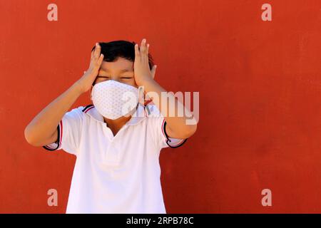 Felice e penoso Latino 8 anni ragazzo che indossa camicia bianca uniforme scolastica e maschera viso per la protezione nella pandemia di Covid-19 al ritorno a scuola Foto Stock