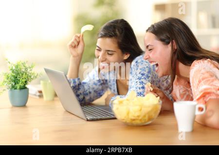 Compagni di stanza felici che ridono ad alta voce guardando video divertenti in un computer portatile mangiando patatine a casa Foto Stock