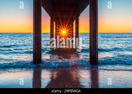 Il sole splende attraverso i piloni del Brighton Jetty mentre si guarda dalla spiaggia al tramonto, nell'Australia meridionale Foto Stock
