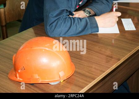Un uomo che lavora come ingegnere con un casco giallo arancio sul tavolo sta studiando, scrivendo in un quaderno in una fabbrica industriale. Foto Stock