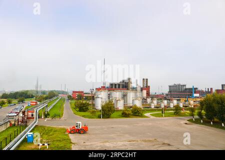 Paesaggio industriale con impianti chimici, tubi e colonne. Di seguito è riportato un trattore arancione. Il fumo proviene dal reattore. Vista panoramica del prodotto di riparazione Foto Stock