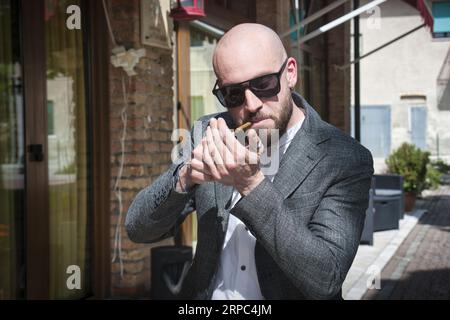 Un giovane con la testa rasata, la barba e gli occhiali da sole, accende un sigaro Foto Stock