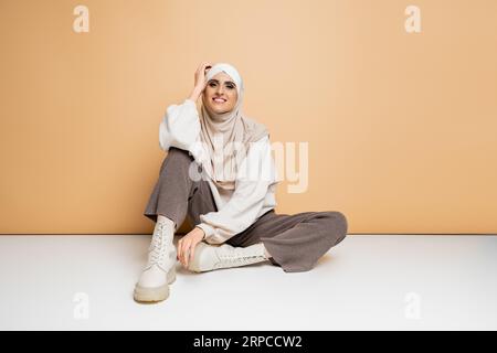 donna musulmana alla moda con hijab, felpa, pantaloni e stivali seduti e sorridenti davanti alla macchina fotografica sul beige Foto Stock