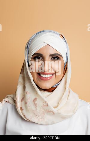 ritratto di affascinante donna musulmana con trucco, in velo di seta sorridente alla macchina fotografica su beige Foto Stock