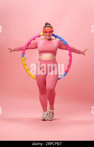 Ritratto a tutta lunghezza di una giovane donna attiva in taglia extra, con top rosa e zampe che fa esercizi con hula hoop, sfondo isolato in studio. Foto Stock