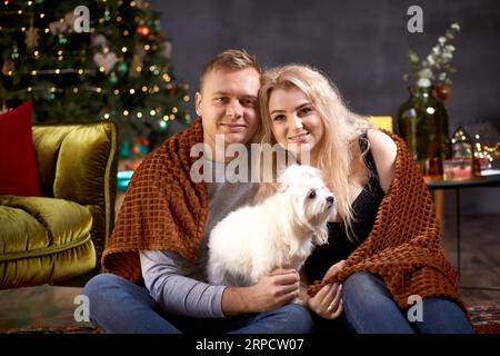 Una coppia romantica e incantevole con il loro cane vicino all'albero di Natale, in un ambiente accogliente ed estetico. Sincero, vero momento. Buon umore e divertimento Foto Stock
