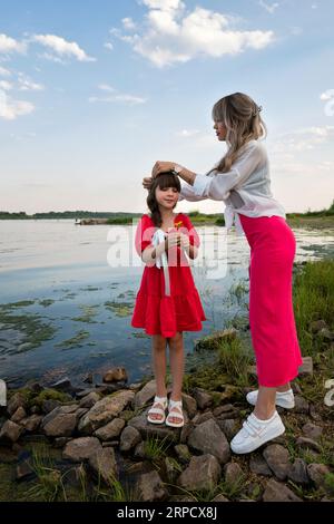 Mamma e figlia stanno camminando vicino al lago o allo stagno. Madre che fissa i capelli della bambina Foto Stock