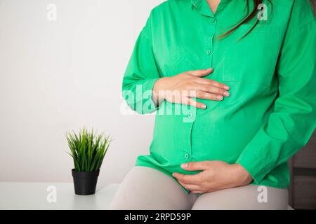 Una ragazza incinta in una camicetta verde le tiene lo stomaco, che ha dolore e gonfiore allo stomaco. Disagio e flatulenza nelle donne in gravidanza. Foto Stock