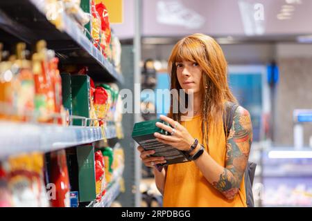 Giovane donna caucasica abbronzata con tatuaggio tiene una confezione di prodotto e sorride. Supermercato in background. Shopping e consumismo. Foto Stock