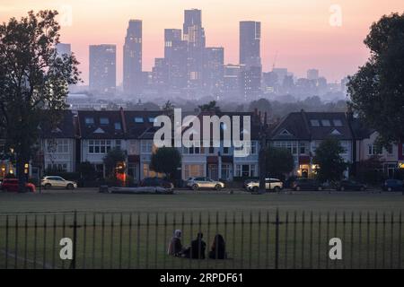 Tre amici si riuniscono per vedere l'ultima luce estiva che svanisce con un punto di vista di case d'epoca terrazzate e in lontananza, il crescente sviluppo a Nine Elms a Battersea, visto da Ruskin Park, uno spazio verde a sud di Londra a Lambeth, il 3 settembre 2023, a Londra, in Inghilterra. Foto Stock