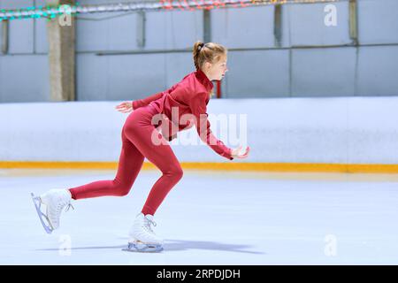 Ragazza adolescente in abbigliamento sportivo rosso, pattinatore di figura in movimento sul ghiaccio, allenandosi nell'arena di pattinaggio, facendo esercizi. Arte Foto Stock