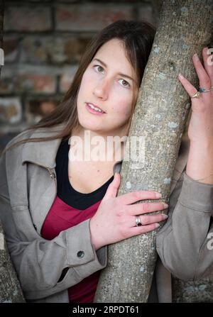 Una donna bruna con camicia rossa e giacca grigia appoggiata a un albero Foto Stock