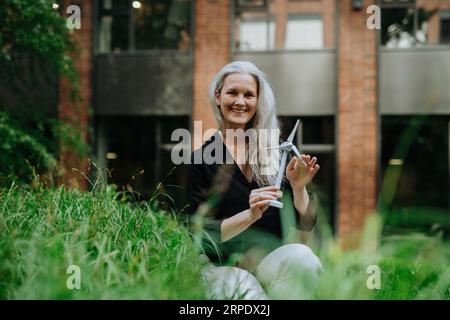 Ritratto di una bella donna con i capelli grigi seduta sull'erba nel parco cittadino, che regge un modello di turbina eolica. Foto Stock