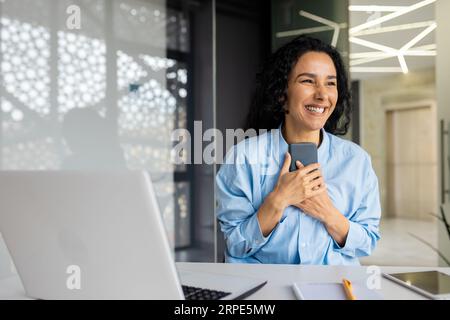 Donna d'affari felice e di successo ha ricevuto una notifica online con buone notizie sul suo telefono, donna ispanica che celebra i buoni risultati e i risultati online, utilizzando l'app sullo smartphone all'interno dell'ufficio. Foto Stock