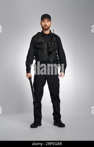 Agente sicuro delle forze dell'ordine in uniforme nera in piedi con la bacchetta in studio. Vista frontale del poliziotto barbuto che tiene il tronco e guarda la fotocamera, su sfondo grigio. Concetto di equipaggiamento della polizia. Foto Stock