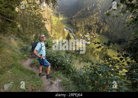 Un uomo dai capelli grigi con zaino e bastoncini si erge su un sentiero che sovrasta una splendida vista di una valle di montagna illuminata dal sole, l'Austria Foto Stock