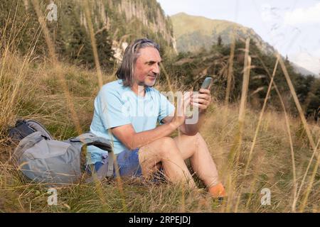 Un uomo a pelo lungo dai capelli grigi con uno zaino e uno smartphone siede nell'erba sullo sfondo delle montagne Foto Stock