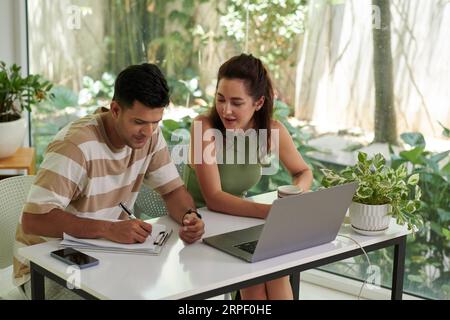 Due giovani colleghi interculturali che organizzano il lavoro o analizzano i dati online mentre prendono appunti sui documenti durante la discussione Foto Stock