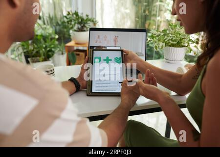 Giovane coppia con tablet rispondendo alle domande di foglio di lavoro online sulla salute mentre l'uomo con stilo mettendo zecche contro sì o nessuna variante Foto Stock