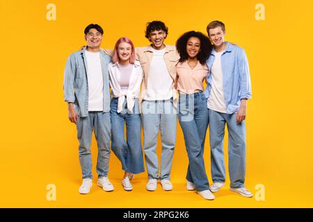 Ritratto di amici felici e diversi che si abbracciano e sorridono davanti alla fotocamera, posare su sfondo giallo, a tutta lunghezza Foto Stock