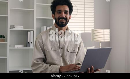 Uomo d'affari indiano musulmano che lavora da casa ufficio lavoro remoto sorridendo arabo barbuto ragazzo 30s freelancer stand con laptop digitazione progetto aziendale Foto Stock