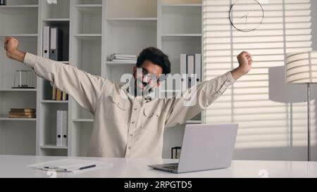 Uomo d'affari arabo stanco uomo musulmano indiano che lavora in remoto felice ragazzo maschio soddisfatto seduto alla scrivania dell'ufficio allungare i muscoli del corpo alleviare la tensione dopo Foto Stock