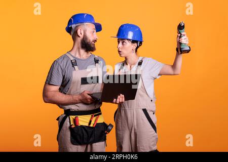 Giovani collaboratori in studio che tengono un computer portatile, stanno cercando di costruire un progetto per un nuovo progetto di ristrutturazione. I rinnovatori che indossano l'uniforme da costruzione prendono decisioni insieme come team. Foto Stock