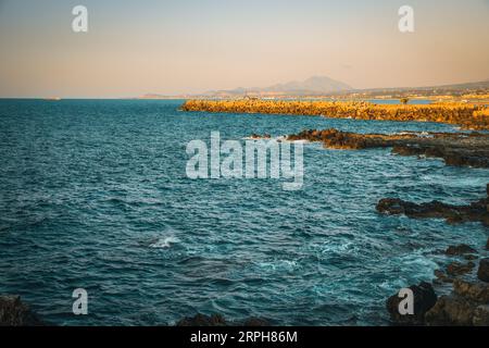 Scopri l'incanto del crepuscolo su una spiaggia rocciosa a Rethymno. Sullo sfondo di maestose montagne e con una nave che scivola sul mare Foto Stock