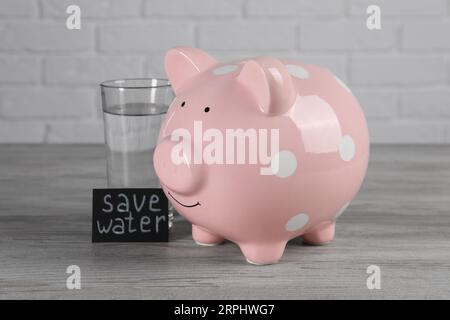 Scheda con frase Salva acqua. Salvadanaio e bicchiere di bevanda su un tavolo di legno Foto Stock