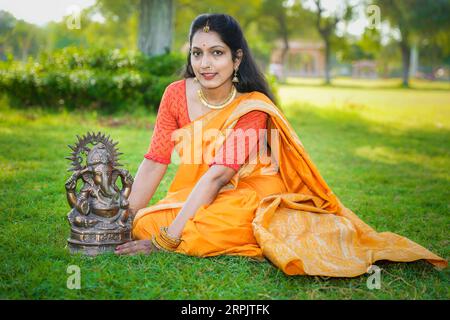 Felice giovane donna indiana che indossa il sari seduta insieme all'idolo di lord Ganesha all'aperto nel parco che celebra il festival di Ganesh Chaturthi. Foto Stock