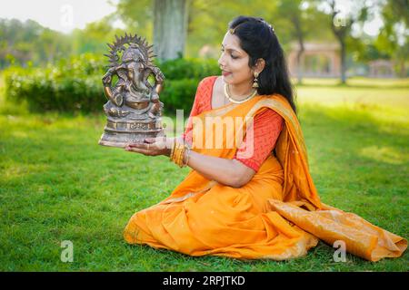 Giovane donna indiana felice che indossa il sari seduto e tiene l'idolo del signore Ganesha all'aperto nel parco che celebra il festival Ganesh Chaturthi. Foto Stock