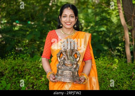 Giovane donna indiana felice che indossa sari che tiene l'idolo del signore Ganesha all'aperto nel parco che celebra il festival Ganesh Chaturthi. Foto Stock