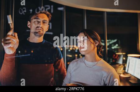 Incontro d'affari a tarda notte con due professionisti che fanno brainstorming di idee, discutono di strategie e collaborano in un ufficio moderno con pareti di vetro. Foto Stock