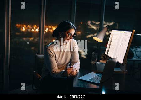 donna d'affari che partecipa a una riunione d'ufficio a tarda notte, utilizza il laptop per una videochiamata. Discute un progetto con i colleghi, mostrando dedizione Foto Stock