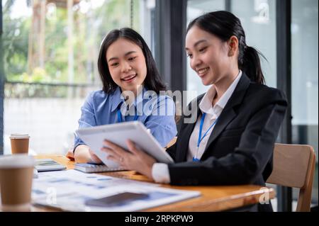 Due giovani donne d'affari asiatiche belle e felici condividono le loro idee di progetto, discutono di lavoro e usano un tablet digitale insieme a un tavolo in Foto Stock