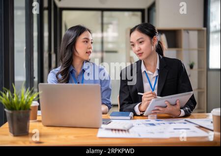Due giovani donne d'affari asiatiche focalizzate o contabili donne stanno discutendo di lavoro, esaminando i dati finanziari e lavorando sui rapporti in ufficio Foto Stock