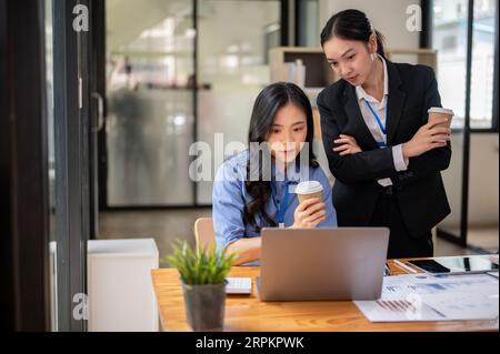 Due donne d'affari asiatiche millenarie e sicure di sé stanno utilizzando un notebook insieme, pianificando e lavorando a un progetto sul laptop in ufficio. Foto Stock