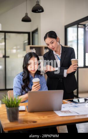 Due donne d'affari asiatiche millenarie, professionali e belle, usano un notebook insieme, lavorano ed esaminano un progetto sul laptop in ufficio Foto Stock