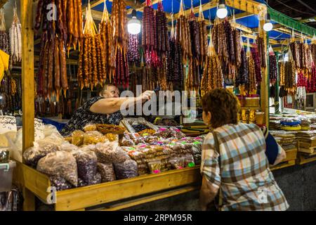 Un commerciante del mercato indica con un coltello strisce di churchkhela (georgiano ჩურჩხელა) appese sopra il suo banco nel mercato coperto dei contadini in via Chavchavadze a Telavi, Georgia. Un cliente con una camicia a quadri e una borsa a tracolla si trova davanti al banco. La dolcezza pende dalle travi di legno in arancione, marrone, rosso e viola. Noci e frutta secca in sacchi sono disposte sul bancone, con barattoli di conserve a destra. Per preparare questo dolce tradizionale georgiano, vengono infilate noci o nocciole su uno spago. Le strisce vengono immerse più volte nel pelamushi, succo d'uva bollito e addensato. Foto Stock