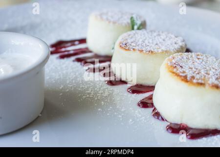 Pancake al formaggio cottage con marmellata di ciliegie e panna acida. Sciroppo alla vaniglia su piatto bianco. Dessert con formaggio curd dolce. Colazione dolce. Foto Stock