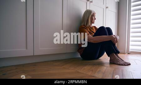 Donna anziana depressa che soffre di cattiva salute mentale seduta sul pavimento a casa Foto Stock