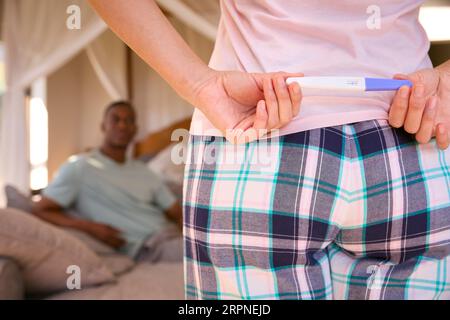 Donna in camera da letto che nasconde il test di gravidanza positivo dietro al partner a sorpresa Foto Stock