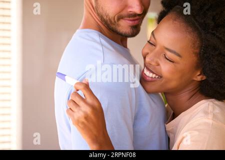 Abbracciare una coppia multi-razziale nella camera da letto a casa celebrando il risultato positivo del test di gravidanza Foto Stock
