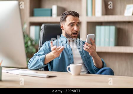Un uomo d'affari scioccato che leggeva un messaggio sullo smartphone mentre era seduto sul posto di lavoro in ufficio Foto Stock